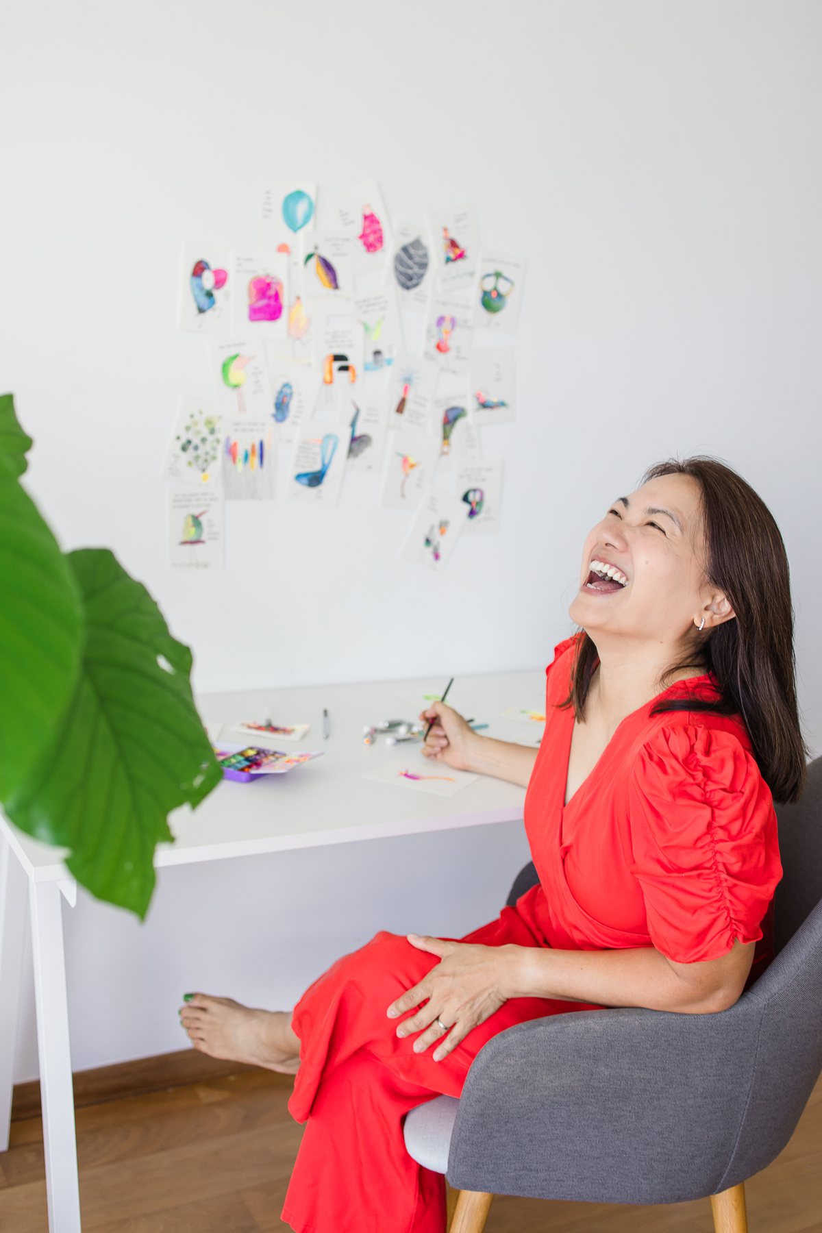Hooi See laughing at her painting desk, her watercolour birds pinned on the wall behind her