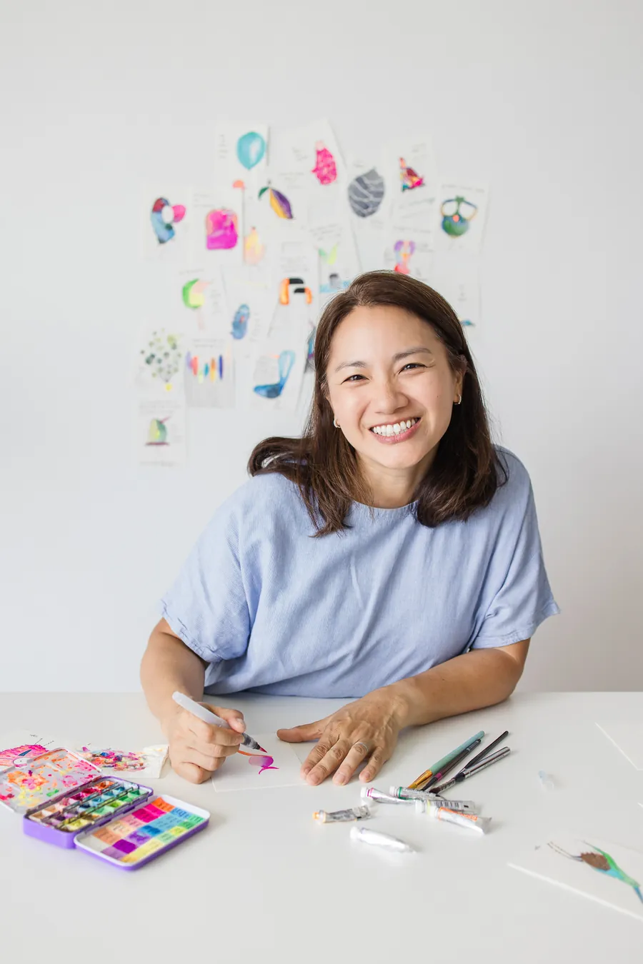 Hooi See at her painting table, smiling at the camera with a brush in her hand, watercolour paints and tubes in front of her and rows of her bird paintings pinned on the wall behind