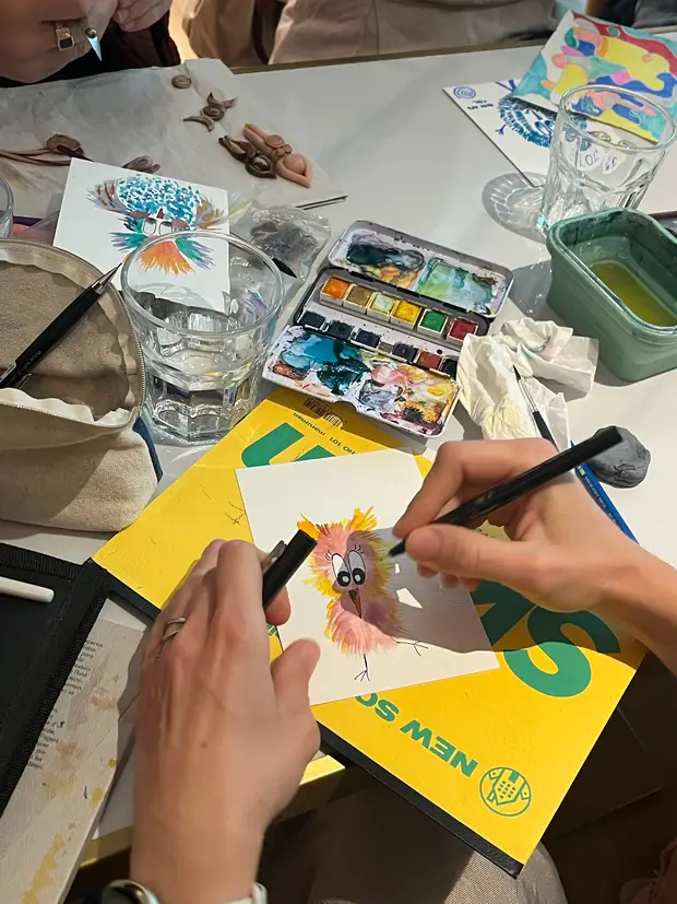 Overhead view of a hand painting a small bird, watercolour box and water jar alongside