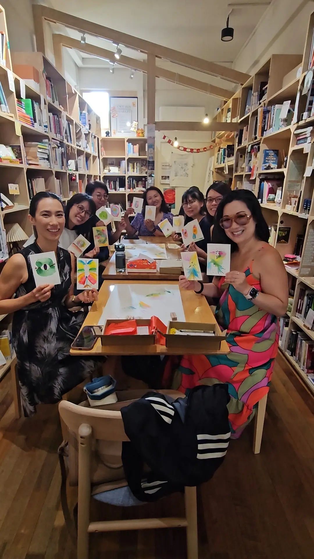 A workshop in a bookshop seen from the end of a long table, shelves down both walls, everyone holding up the bird they painted