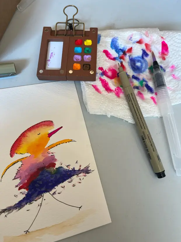 A finished orange and purple watercolour bird beside a paintbox and brush