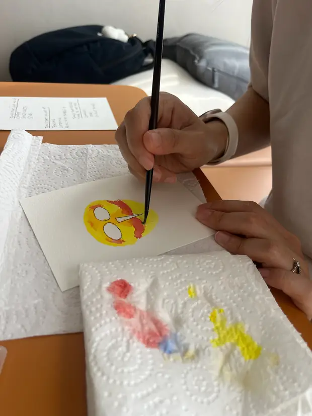 A hand painting a small yellow bird with a brush, colour tests on the paper alongside