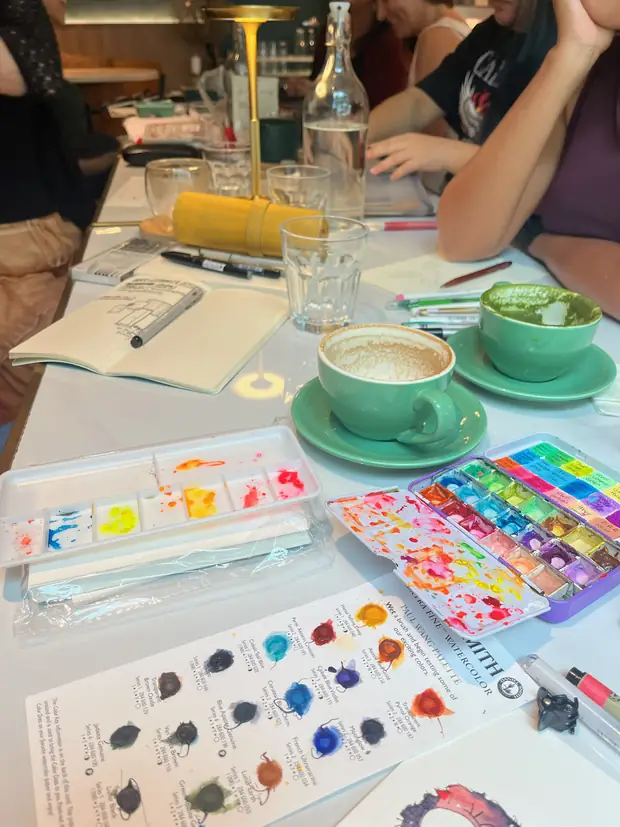A café table laid out with open paintboxes, mixing palettes and cups of coffee