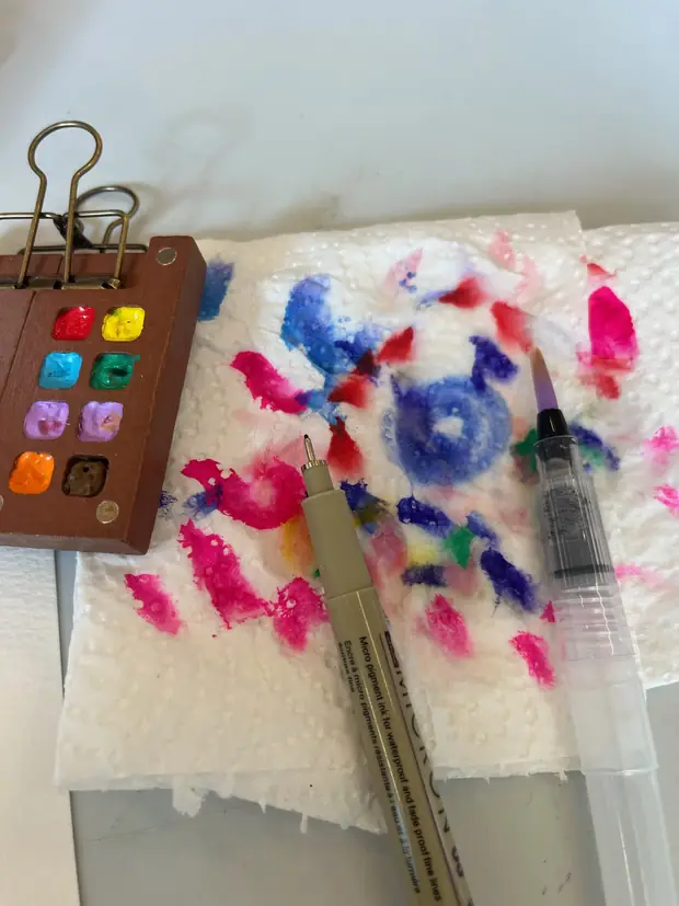 A paintbox and two brushes beside a sheet of pink and blue colour tests