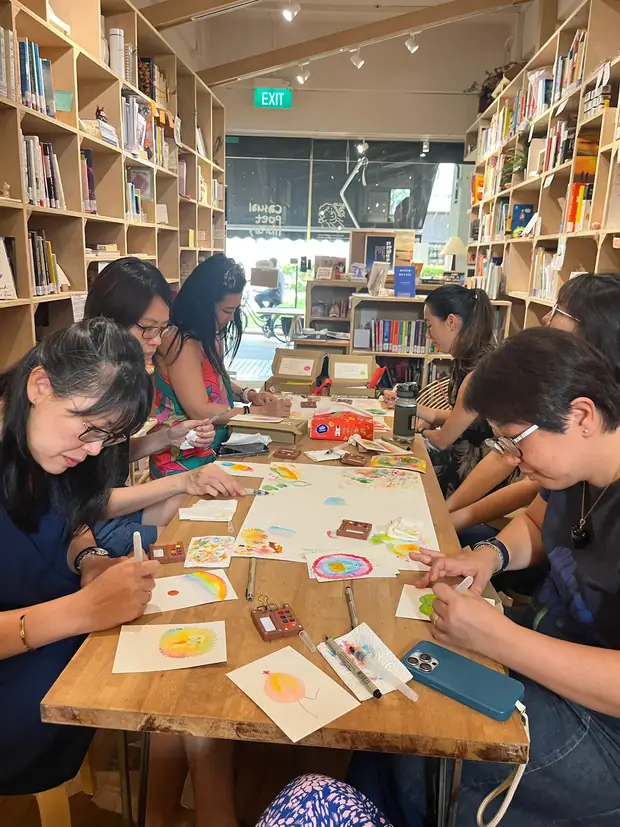 Seven people painting at a long wooden table in a bookshop, shelves to the ceiling on both sides and the shopfront window behind