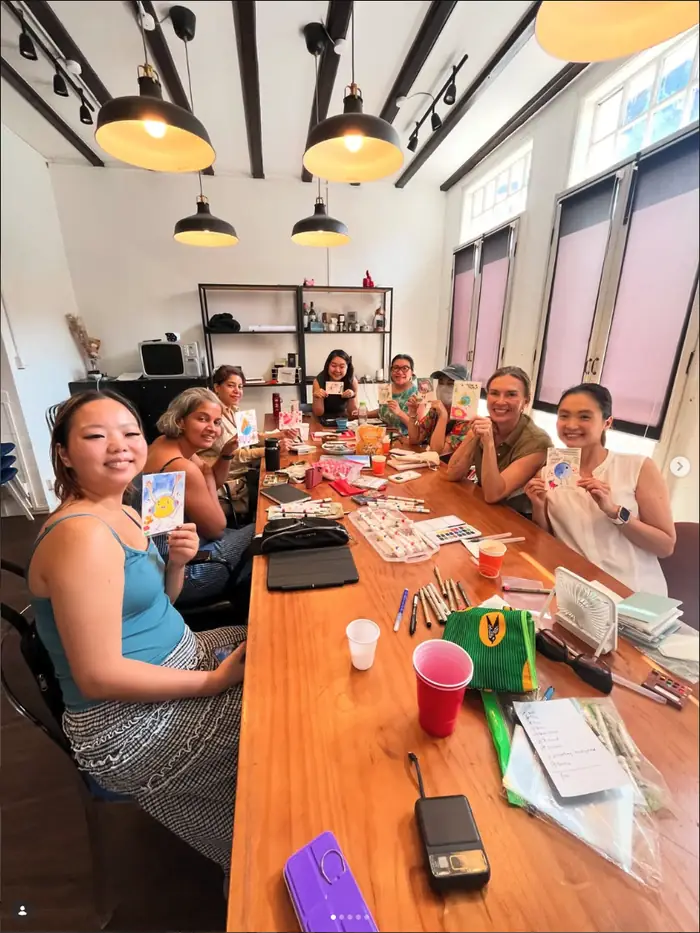 A long shared table under pendant lights, eight people around it holding up the birds they painted